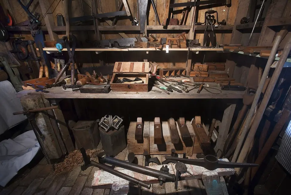 Établi ancien du XIXe siècle encombré de rabots, vilebrequins et outils de menuisier, sous des étagères chargées d'outils en bois.