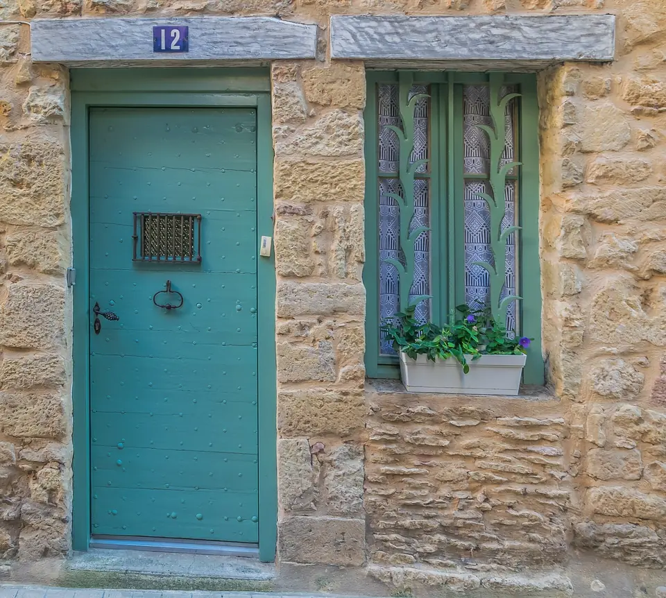 Porte d'entrée en bois peinte en vert d'eau avec judas grillagé, à côté d'une fenêtre fleurie dans un mur en pierre blonde.